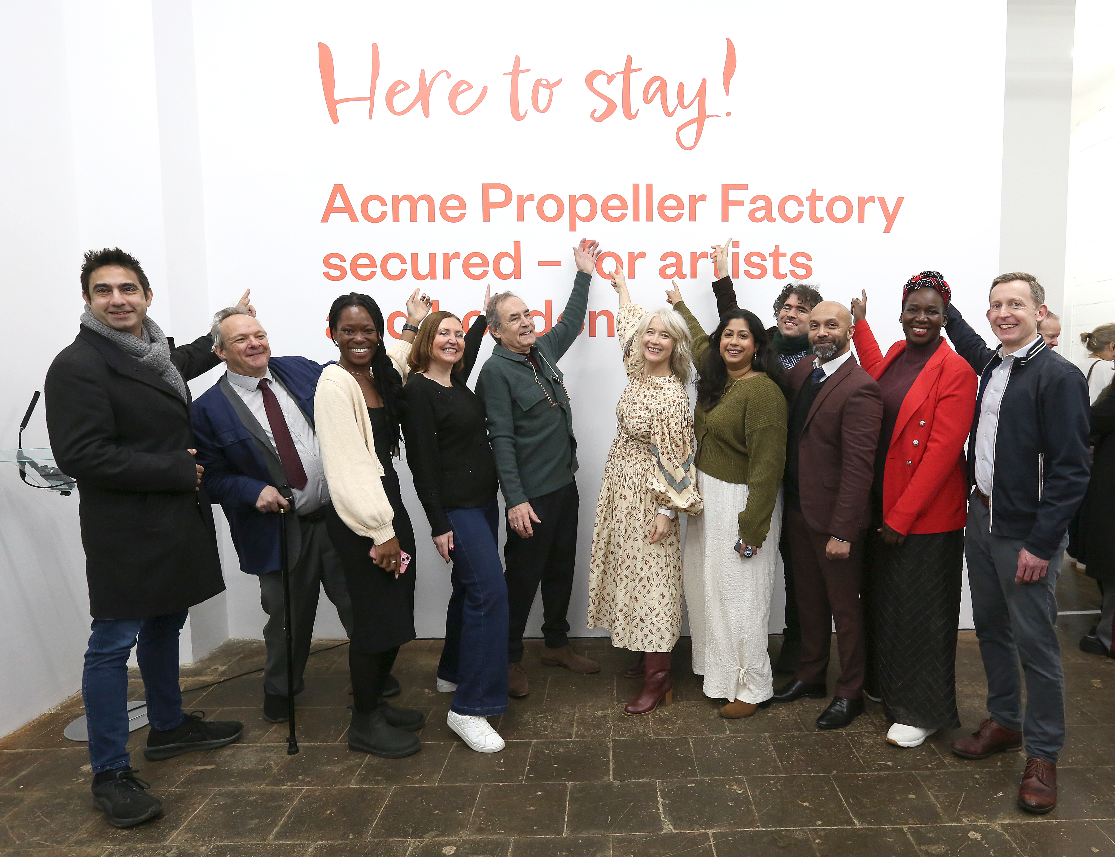 Group of diverse people standing in front of a wall with text about Acme Propeller Factory securing space for artists, celebrating together.