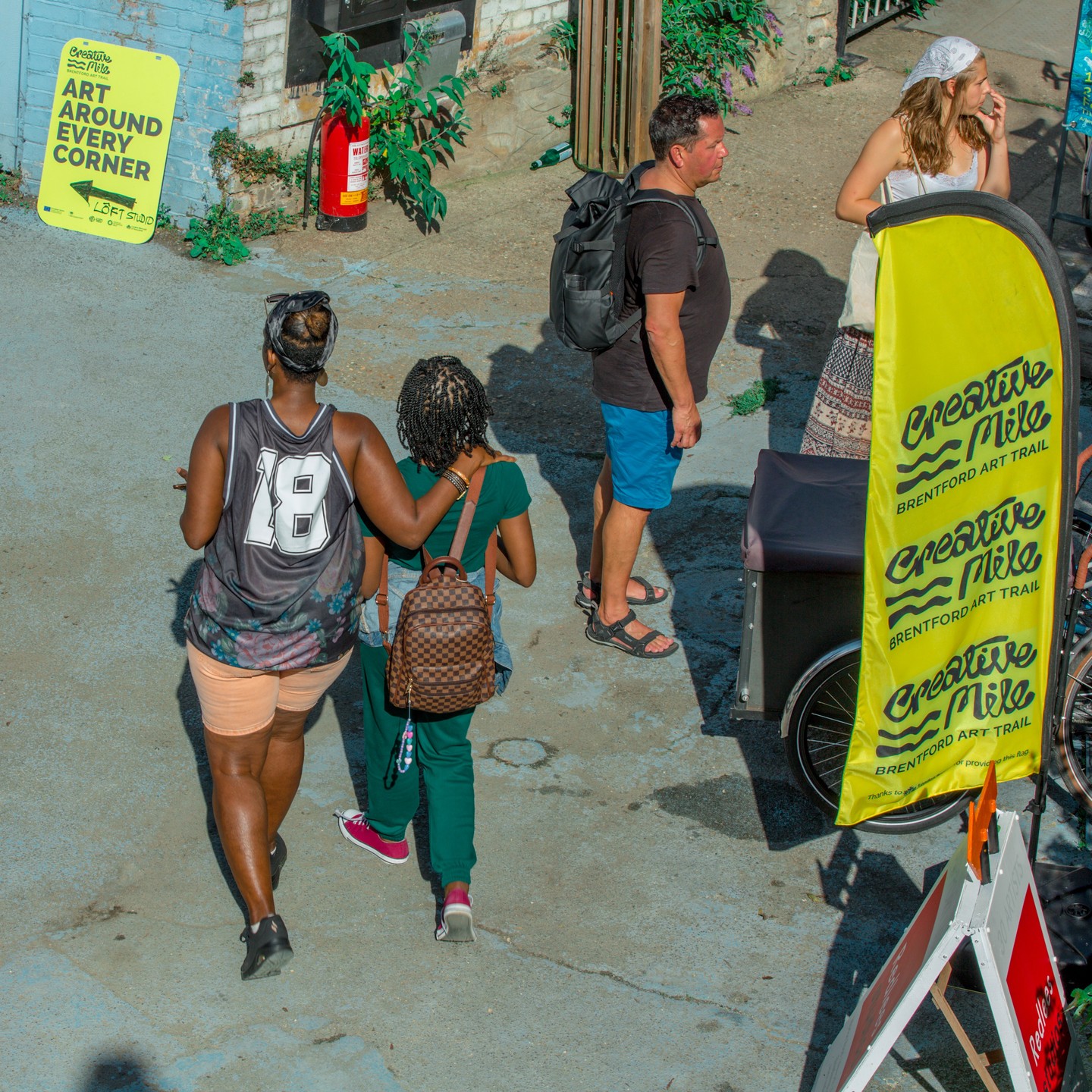 Two women walk arm in arm near a Creative Mile Brentford Art Trail sign on a sunny street corner.