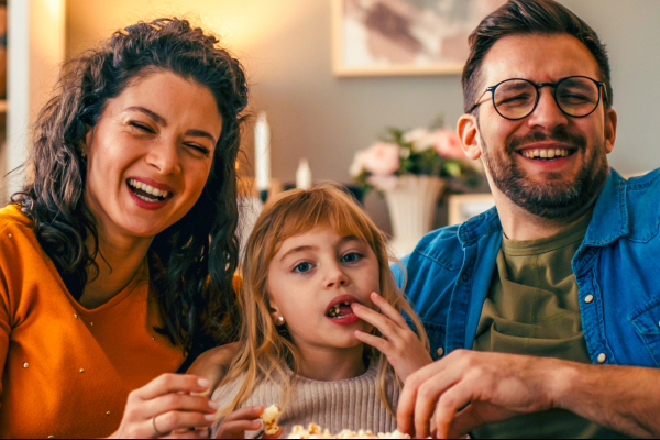 A young couple with child on sofa at home eating popcorn and smiling at the camera