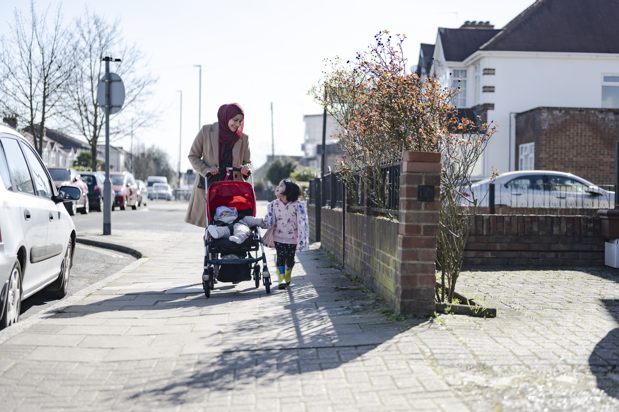 a lady walking down the pavement pushing a young child in a pram with another child walking alongside her