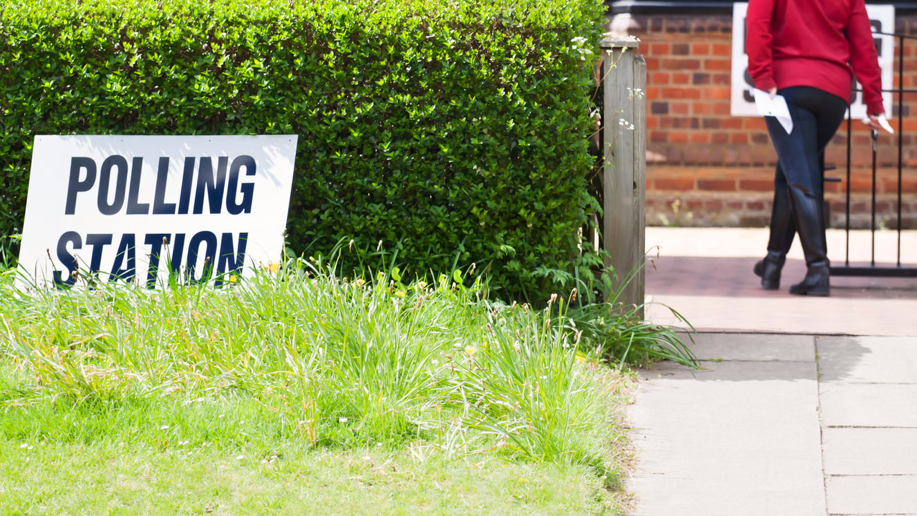A person walking through a gate with a hedge on the left and a sign reading 'Polling Station' in front