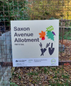 Sign on a wire fence reading ‘Saxon Avenue Allotment’, featuring colorful leaf and gardening tool illustrations.
