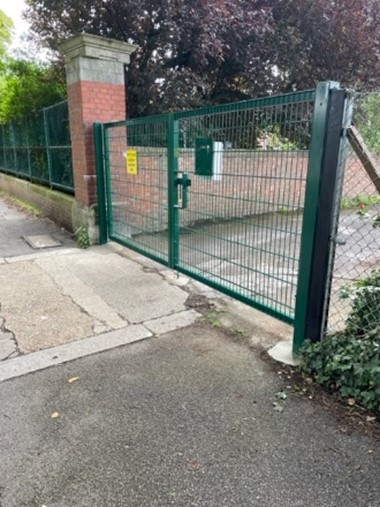 Green metal gate set into a brick wall and fence, opening from a pavement into a secured outdoor area with trees behind.