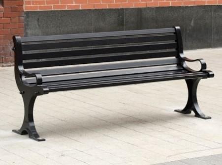 Black metal bench with slatted backrest and seat, positioned on a light grey paved surface against a brick and stone wall.