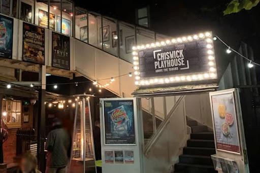 Entrance to Chiswick Playhouse at night with illuminated sign, posters on display, and string lights above a staircase leading inside.