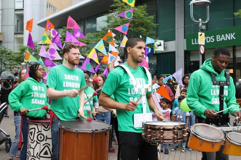 Street parade with drummers in green ‘South London Samba’ shirts, colourful flags in the background, and a crowd watching