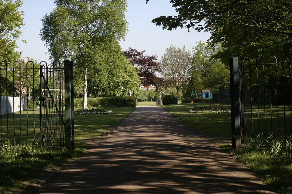 Open park entrance with a paved path leading through green lawns and trees under clear blue sky.