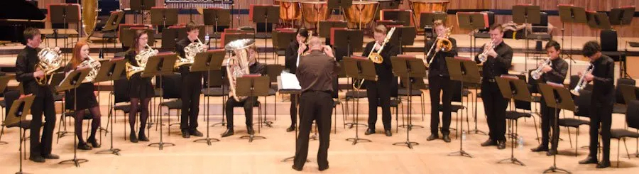 A conductor leads a group of brass musicians performing on stage, with music stands arranged in a semicircle.