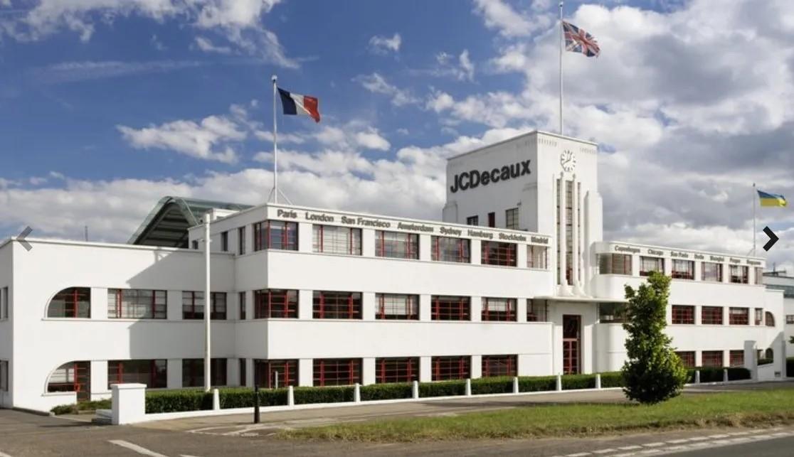 Art Deco-style white building with red-framed windows and a central tower displaying the JCDecaux logo, topped with a clock.