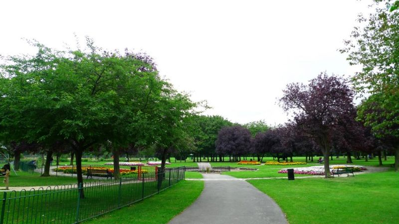 Park with paved paths, green lawns, and colourful flowerbeds surrounded by trees under an overcast sky.