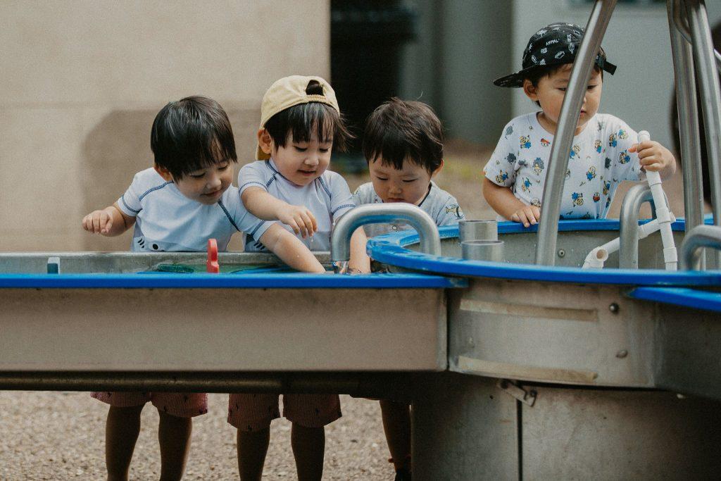 Four young children standing at a water play table outdoors, interacting with flowing water and metal fixtures.