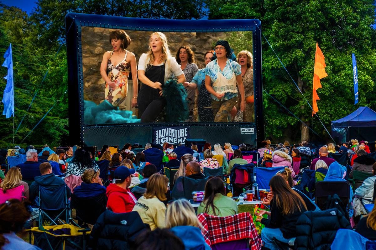 Outdoor cinema event with a large screen showing a movie scene, surrounded by trees and colourful flags, as a seated audience watches on folding chairs and blankets.