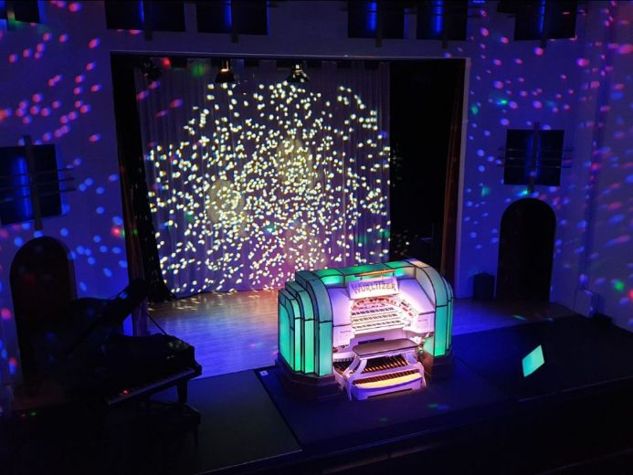 Illuminated Wurlitzer organ on a stage with colourful disco-style light projections on the walls and backdrop.