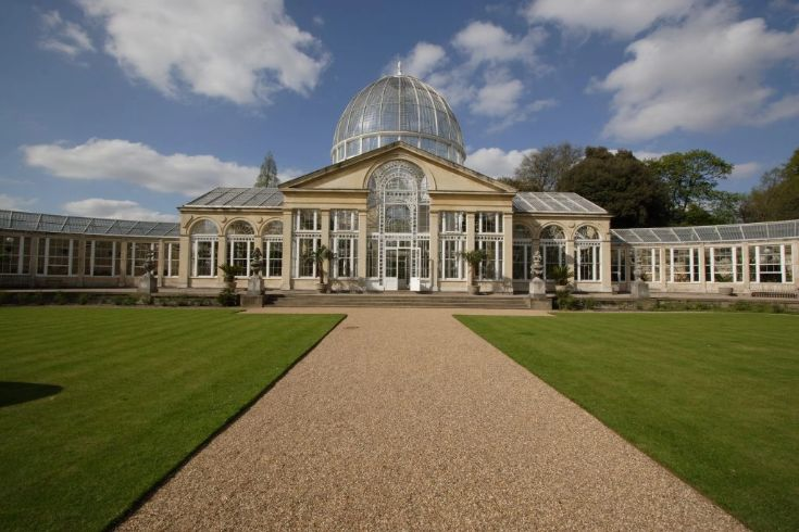 Grand glass conservatory with a central domed roof and arched windows, set behind a gravel path and manicured lawns under a partly cloudy sky.