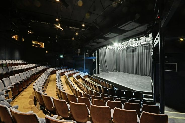 Theatre with curved rows of brown seats facing a stage with black curtains and overhead lighting rigs.